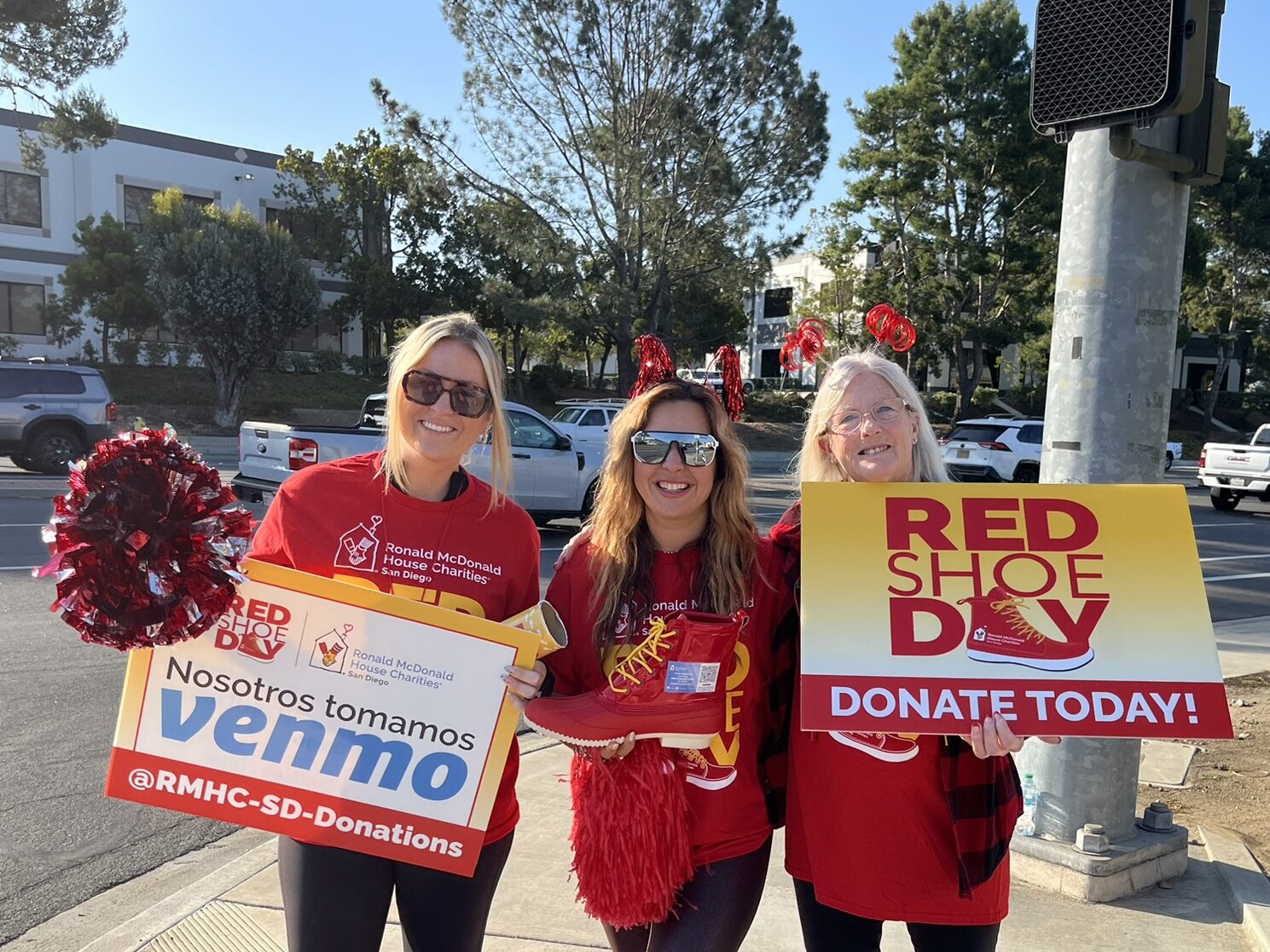 Happy Feet for a Happy House team members in red hold Red Shoe Day donation signs and an oversized red shoe at a San Diego intersection.