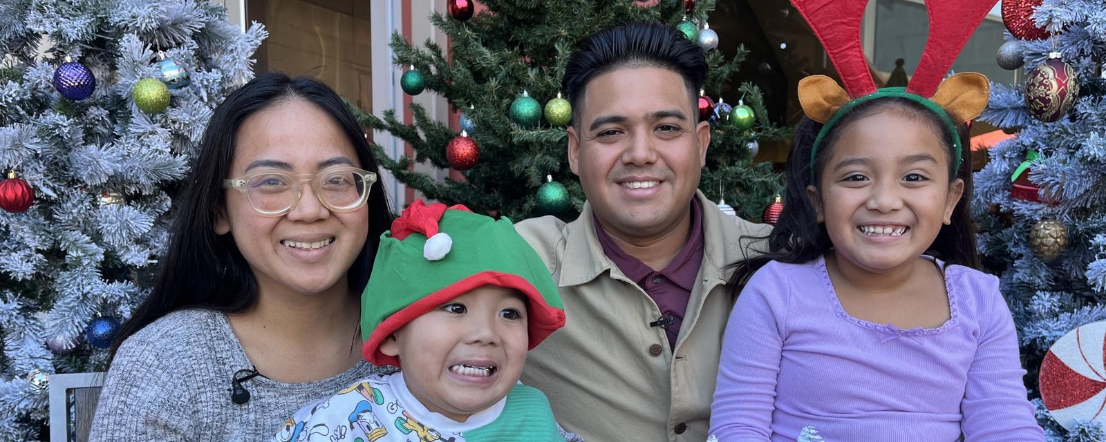 A family of four smiles in front of decorated Christmas trees; the children wear an elf hat and reindeer antlers